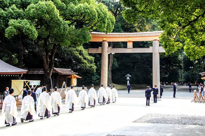 Priests in white hakama walk single file toward a large toori, a gate seen at Japanese shrines. It appears to be summer and the shrine grounds are full and green.