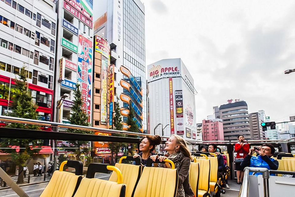Two women in the center of the image take in the sights with other passengers on the second story of an open air bus. Around them are large office and residential buildings somewhere in the east of downtown Tokyo.